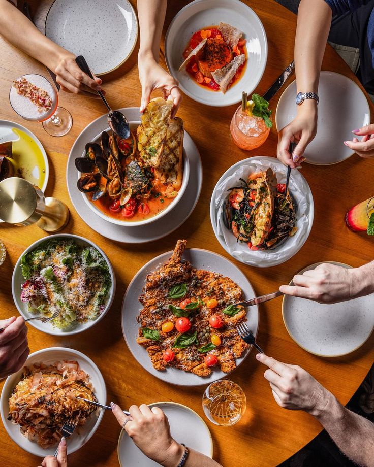 Overhead flatlay of brunch dishes with hands reaching across a wooden table filled with seafood, salads, and sides