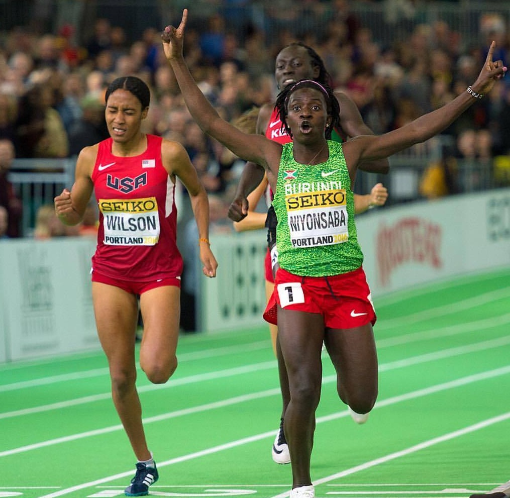 Two female sprinters racing on an indoor track, with the leading runner raising her arms