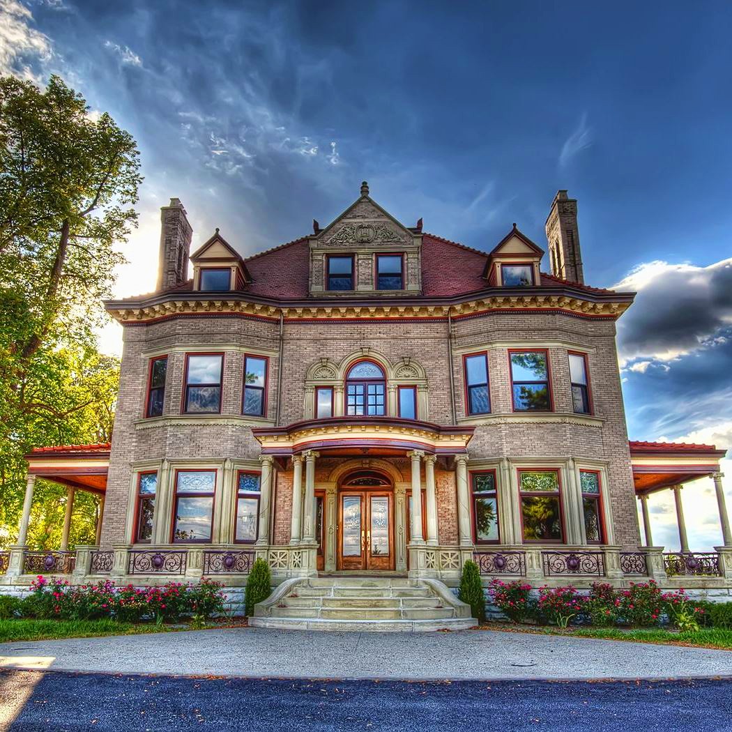 Three-story Victorian mansion with red brick, ornate windows, front stairs, and blue sky