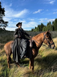 Woman in dark period clothing on horseback in grassy field with forest background