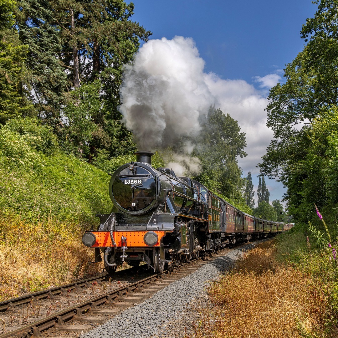 Black vintage steam train with white smoke traveling through green countryside on sunny day