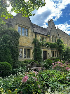 Stone cottage behind a lush flower garden with ivy, trees, and a blue cloudy sky.
