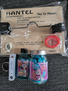 Packaged novelty dumbbell item on a dark surface next to a colorful bottle and a white lighter.