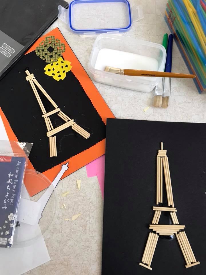 Two matchstick Eiffel Tower models on black paper with craft supplies on a desk