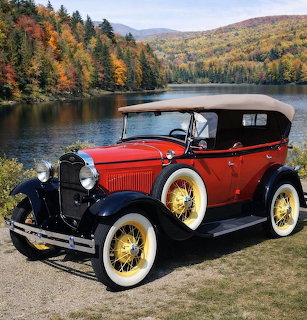 Red and black 1931 Ford Model A Phaeton parked beside a lake with autumn trees and hills