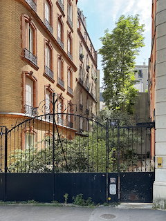 Ornate wrought iron gate opening to a courtyard between Parisian brick apartment buildings with green trees