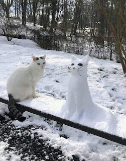 White cat sitting next to a small snowman on a snow-covered wooden deck in winter