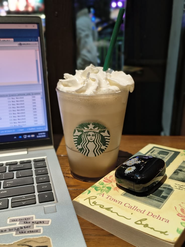 Starbucks Frappuccino with whipped cream next to a laptop and a book on a wooden café table