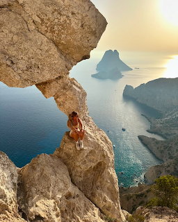 Woman sitting on rocky cliff overlooking blue sea and distant island at sunset