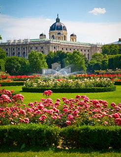 Ornate palace building behind formal garden with red and pink flowerbeds, green lawn, and monument in Vienna