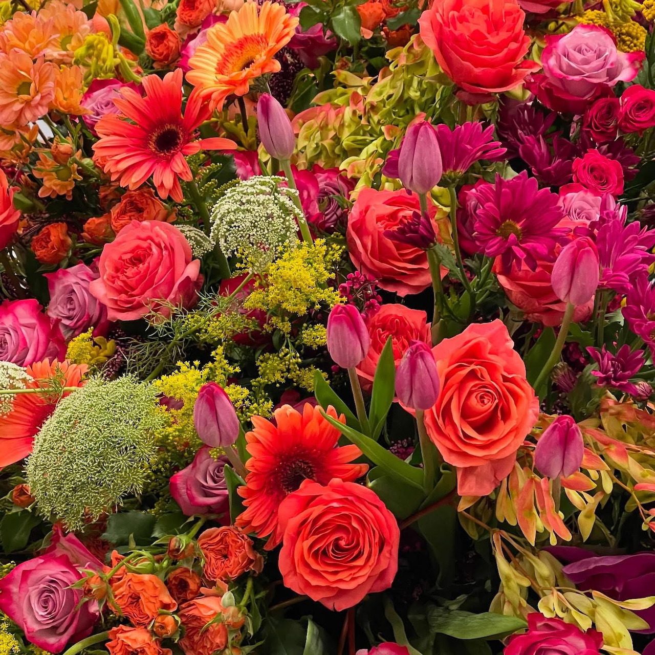 Dense bouquet of pink roses, orange gerberas, purple tulips, and yellow wildflowers with green accents