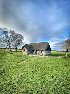 Stone cottage with thatched roof on grassy field with trees and cloudy sky