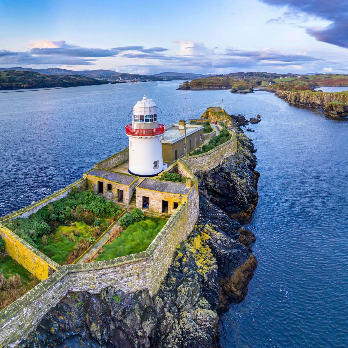 White lighthouse with red band on rocky peninsula surrounded by ocean water with green hills in background