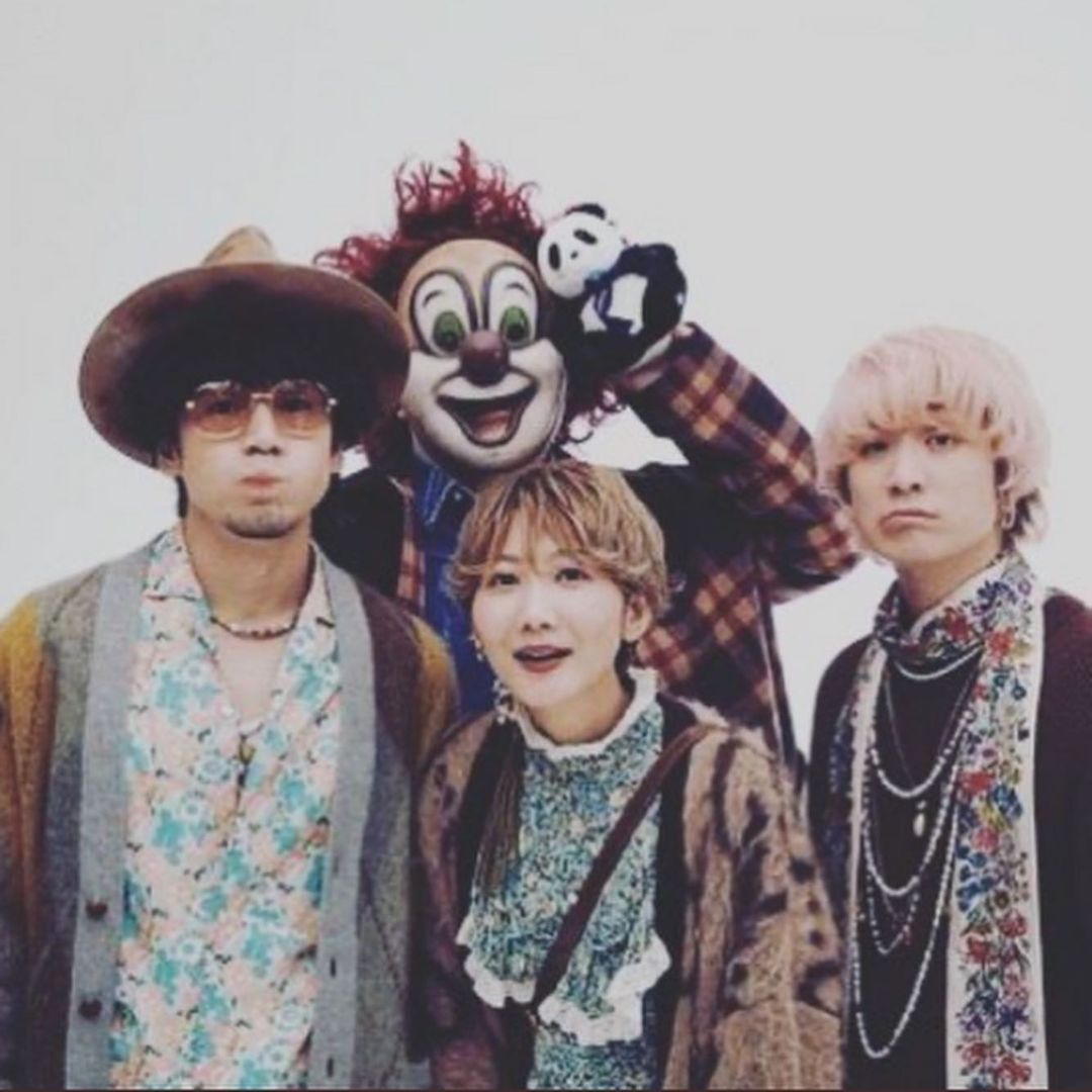 Four band members in colorful eclectic clothing pose in a studio against a white background
