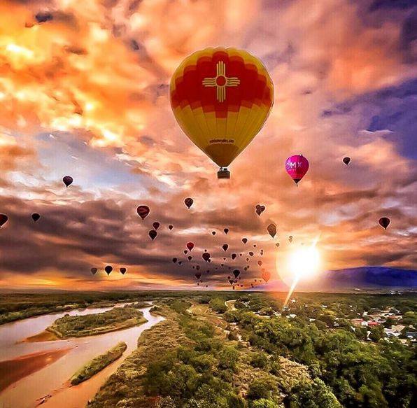Hot air balloons including New Mexico flag balloon floating at sunset over landscape with river