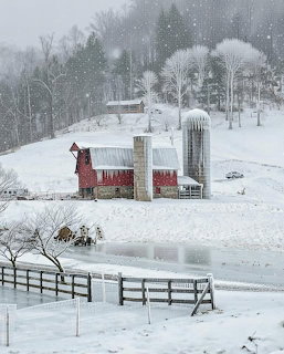 Red barn with stone silos in a snowy winter landscape with falling snow and bare trees