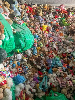 A large pile of colorful stuffed animals and plush toys covering an indoor floor around green bags.