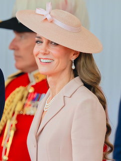Kate Middleton smiling in a beige hat with bow, pearl necklace, and light pink blazer at a formal event