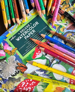 Watercolor paper pad surrounded by colored pencils and crayons on illustrated nature artwork