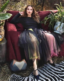 Woman in dark top and colorful tulle skirt sitting on burgundy velvet chair with plants