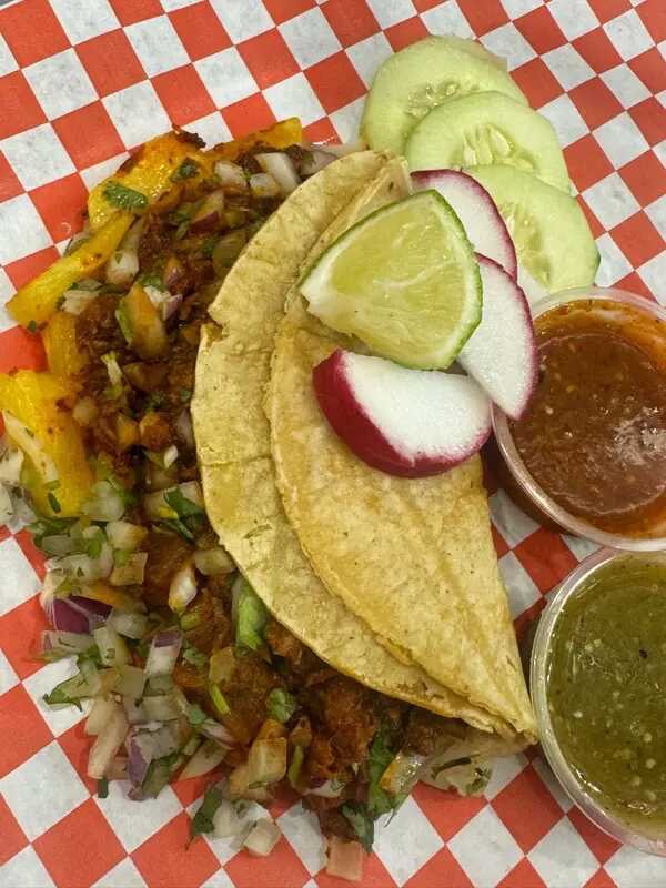 Beef tacos with onion and cilantro, lime, radish, cucumber, and two salsa cups on checkered paper