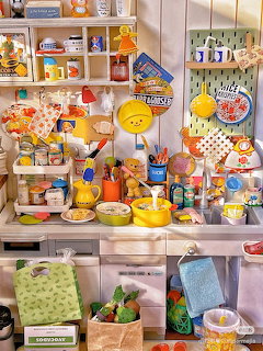 Miniature kitchen shelf display with tiny food items, dishes, and colorful accessories on white shelving