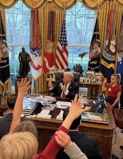 Donald Trump at the Resolute Desk in the Oval Office with children raising their hands