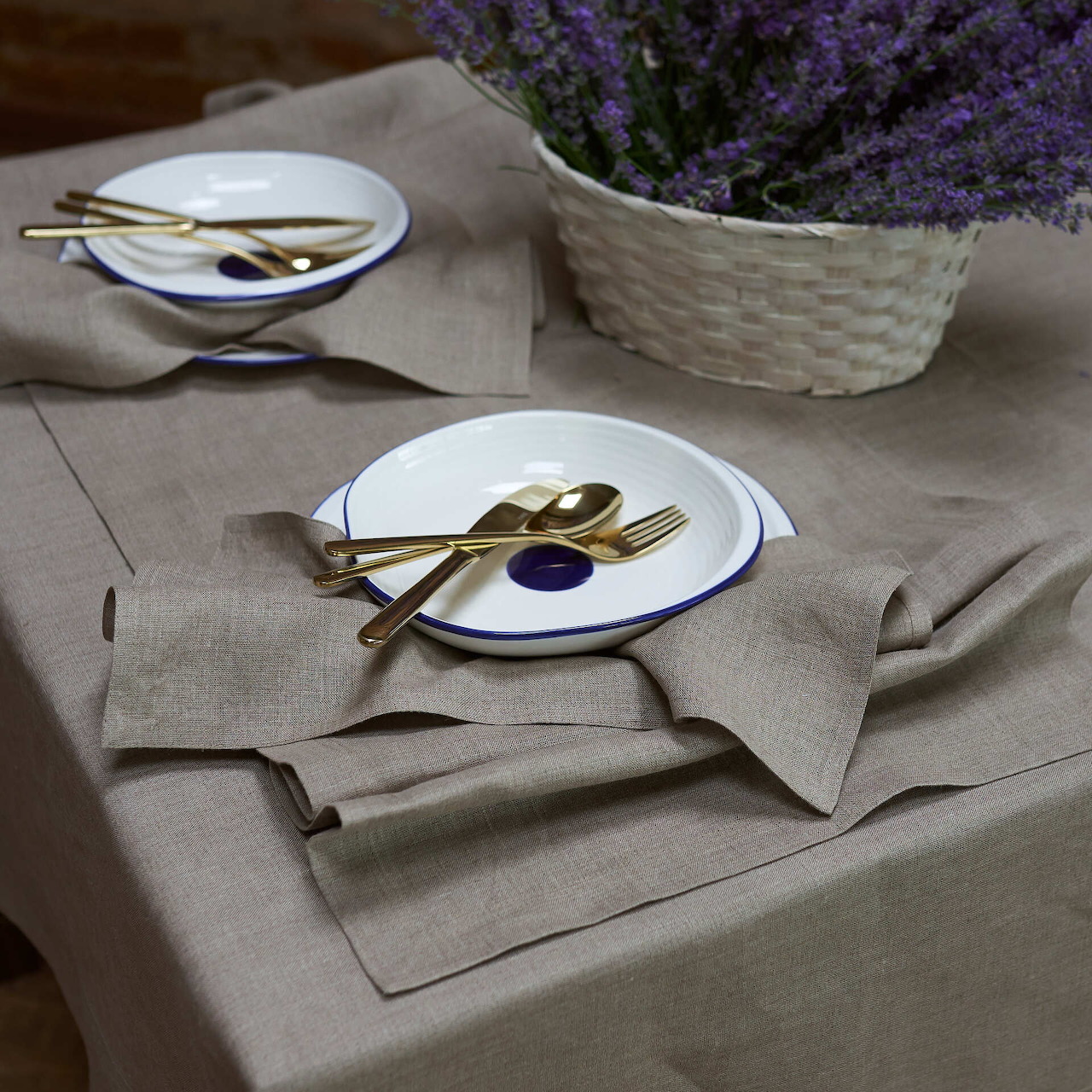 Rustic linen table setting with white plates, gold cutlery, and lavender basket on beige tablecloth