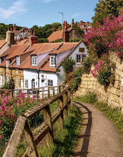 Narrow path with wooden fence alongside stone wall and pink flowers leading to English cottages with red rofs