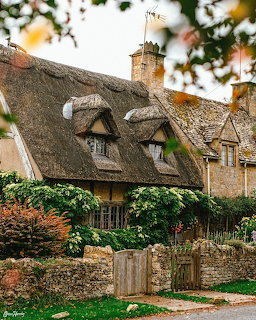 Thatched stone cottage behind a low stone wall and wooden gate with greenery in front
