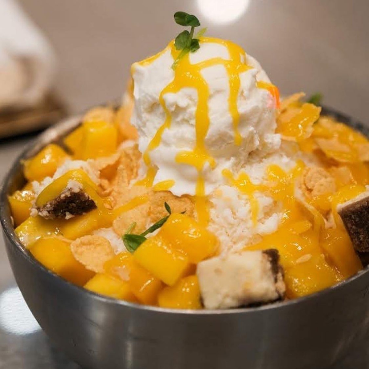 Bowl of mango bingsu topped with whipped cream, mango cubes, sauce, and mint garnish