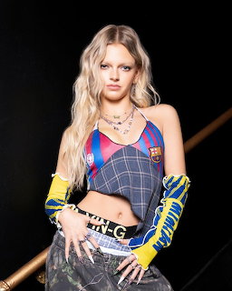 Young blonde woman in layered Barcelona jersey and plaid corset top with yellow gloves posing against dark background