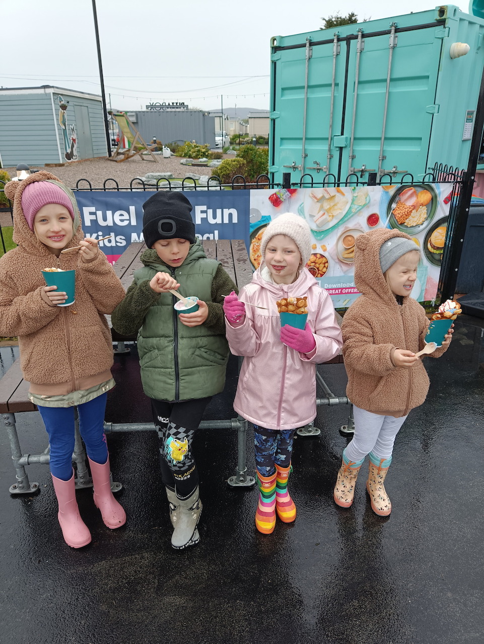 Four children in coats and rain boots holding snack cups outdoors near a blue shipping container