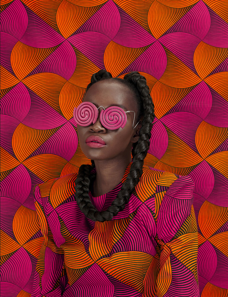 Woman in pink spiral glasses with a bright orange and magenta patterned outfit and matching backdrop