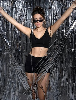 Woman in black crop top and shorts posing with raised arms against silver tinsel backdrop wearing sunglasses