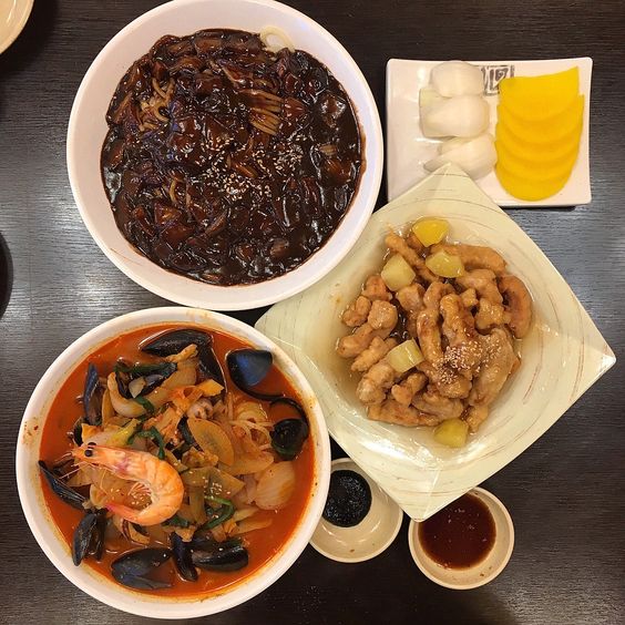 Top-down view of Korean dishes including black bean noodles, seafood soup, sweet and sour pork, and side dishes