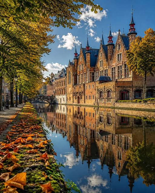 Gothic brick buildings reflected in a calm canal in Bruges with autumn leaves along the walkway
