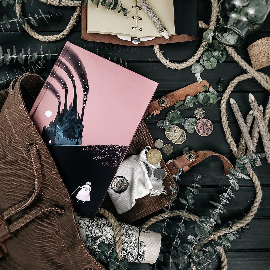 Overhead view of reading setup with book, notebook, coffee, bag, and plants on dark wood table
