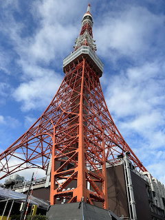 Low-angle view of Tokyo Tower against a blue sky with clouds and buildings below