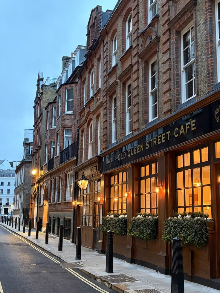 Victorian brick buildings along a London street at night with a warmly lit cafe and green hedges