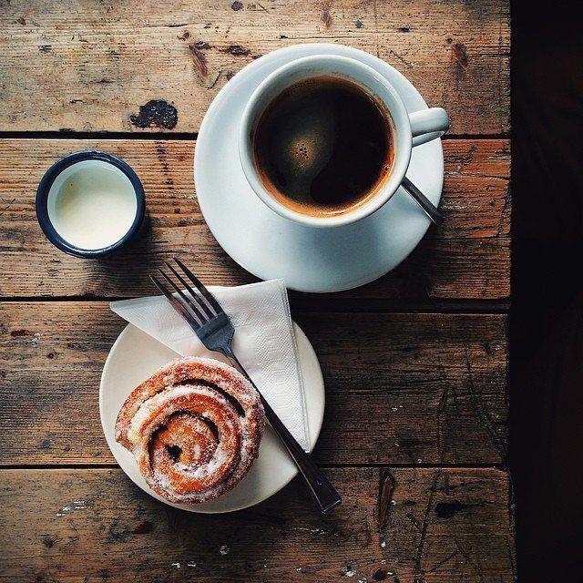 Black coffee with a cinnamon bun, milk pitcher, and fork on a wooden table