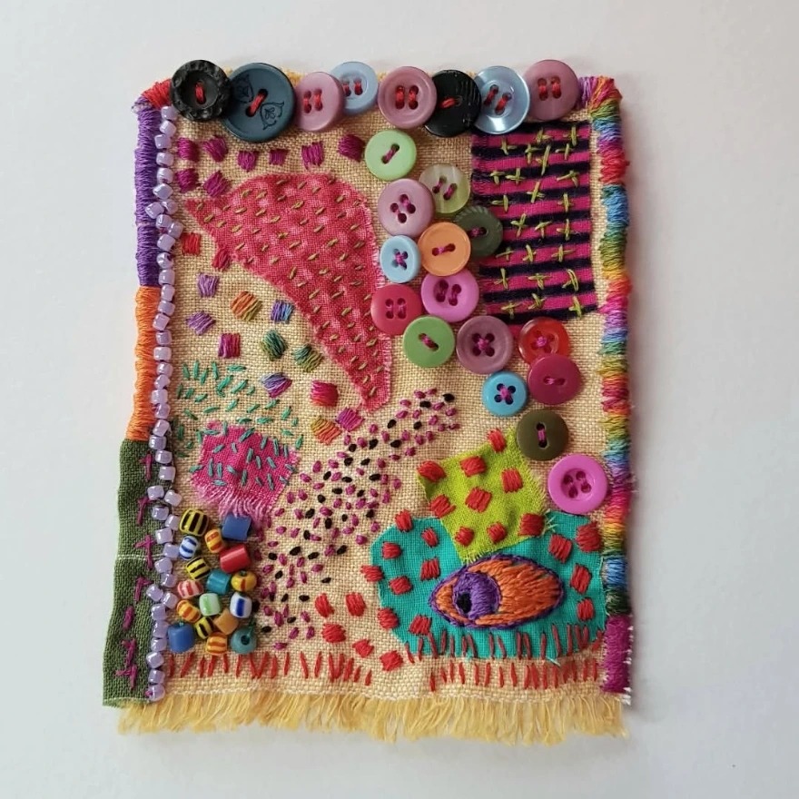 Colorful abstract embroidered fabric panel with buttons, stitching, and fringe hanging on a white wall