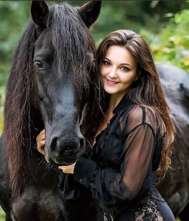 Smiling woman with long brown hair standing next to a black horse outdoors