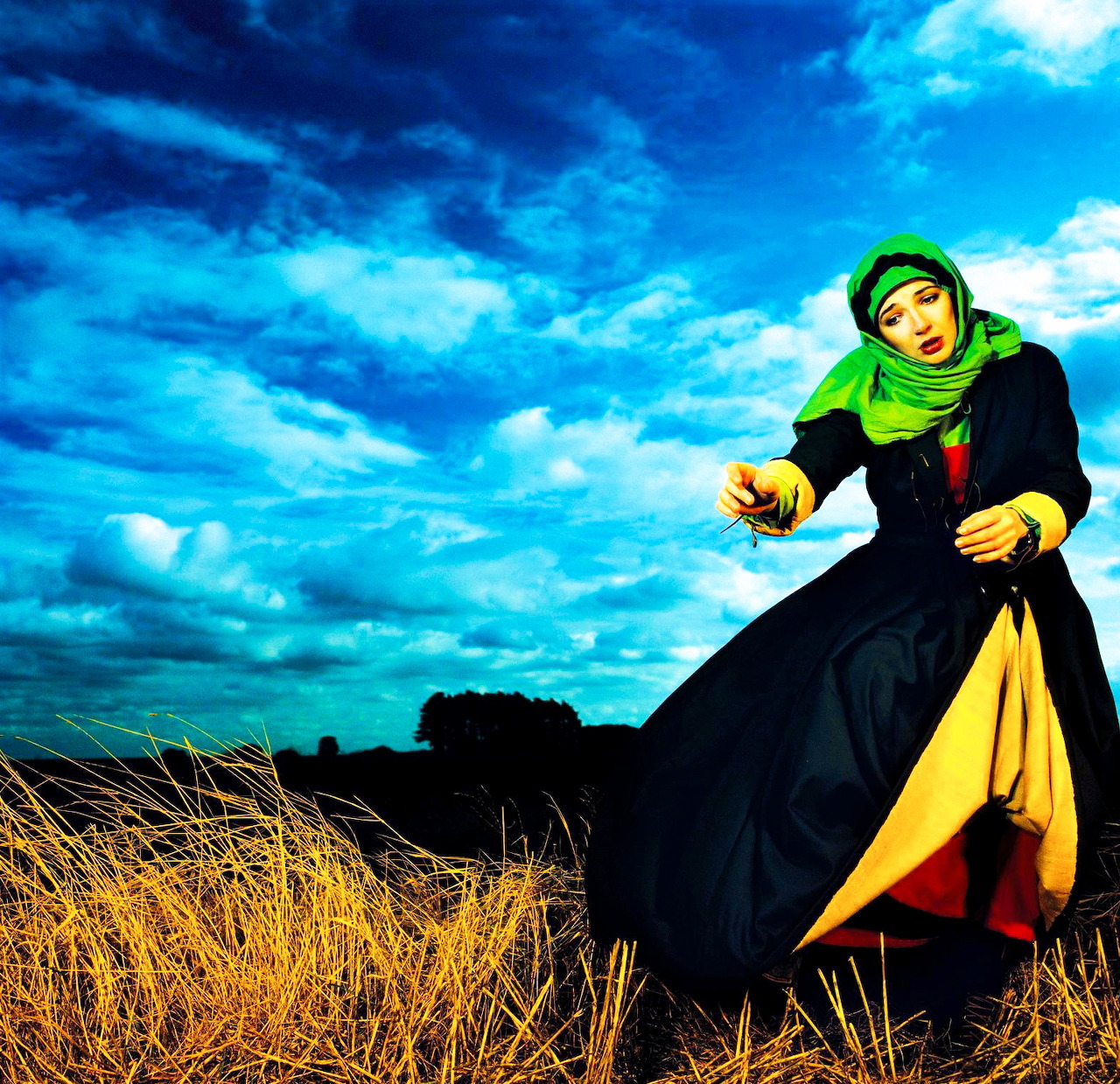 Woman in black coat and green headscarf in windswept field under blue cloudy sky