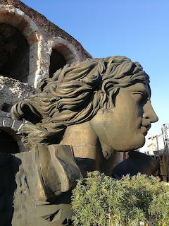 Stone sculpture of woman's head in profile with wavy hair, ancient stone arches visible behind against blue sky