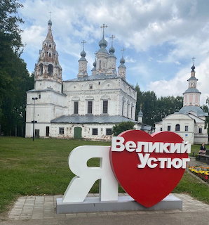White Orthodox church with green domes behind a red heart-shaped I Love Velikiy Ustyug sign in a park