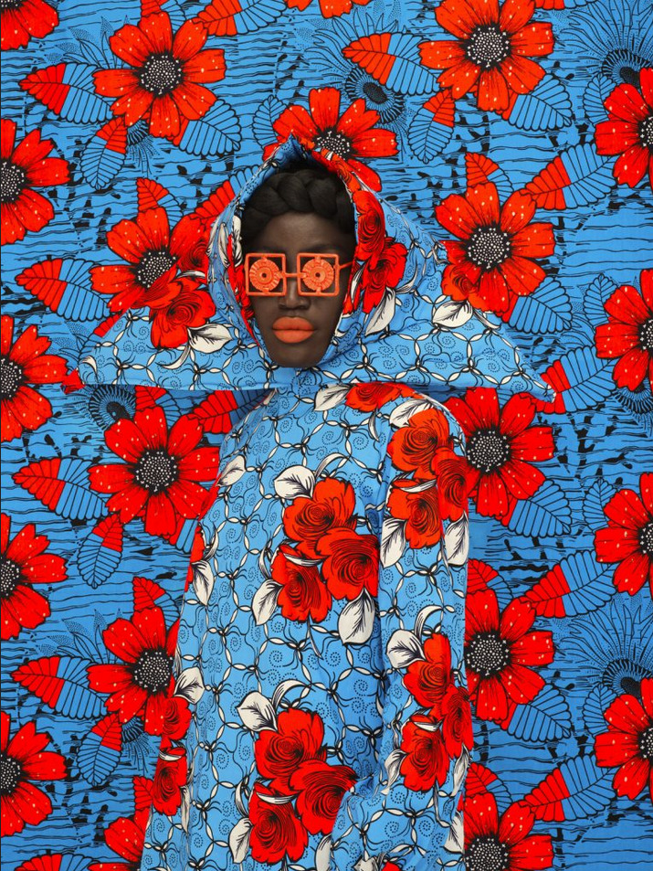 Person blending into blue and red floral African wax print fabric, wearing matching outfit and orange sunglasses