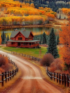 Red log cabin beside a lake surrounded by colorful autumn trees with a dirt road and wooden fences in foreground