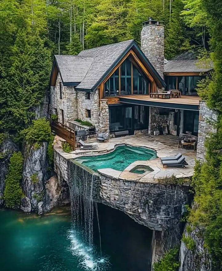 Stone house on a rocky cliff above a turquoise lake, with a pool, waterfall, and forest surroundings
