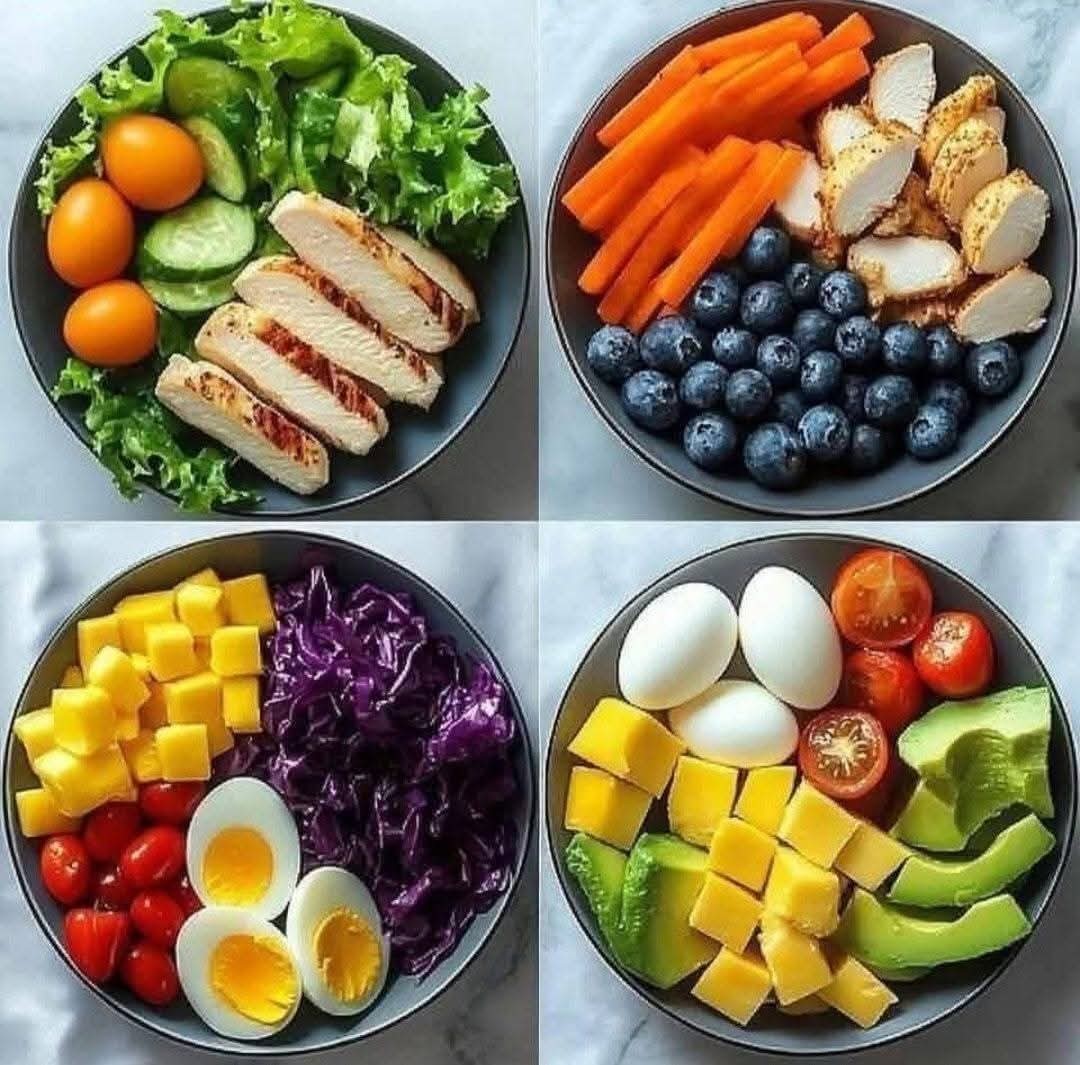 Four circular meal prep bowls with chicken, vegetables, fruits, eggs and greens on marble background